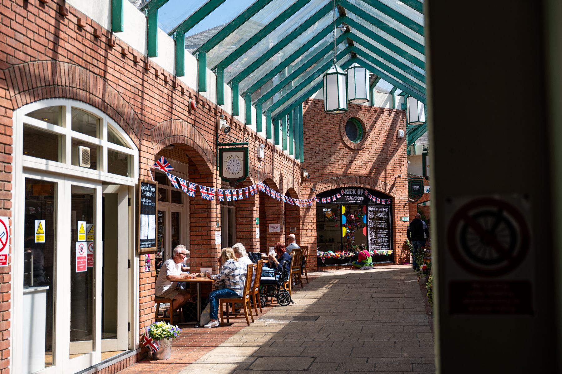 People seated outside cafés in a covered brick-lined shopping arcade with bunting and shopfronts.